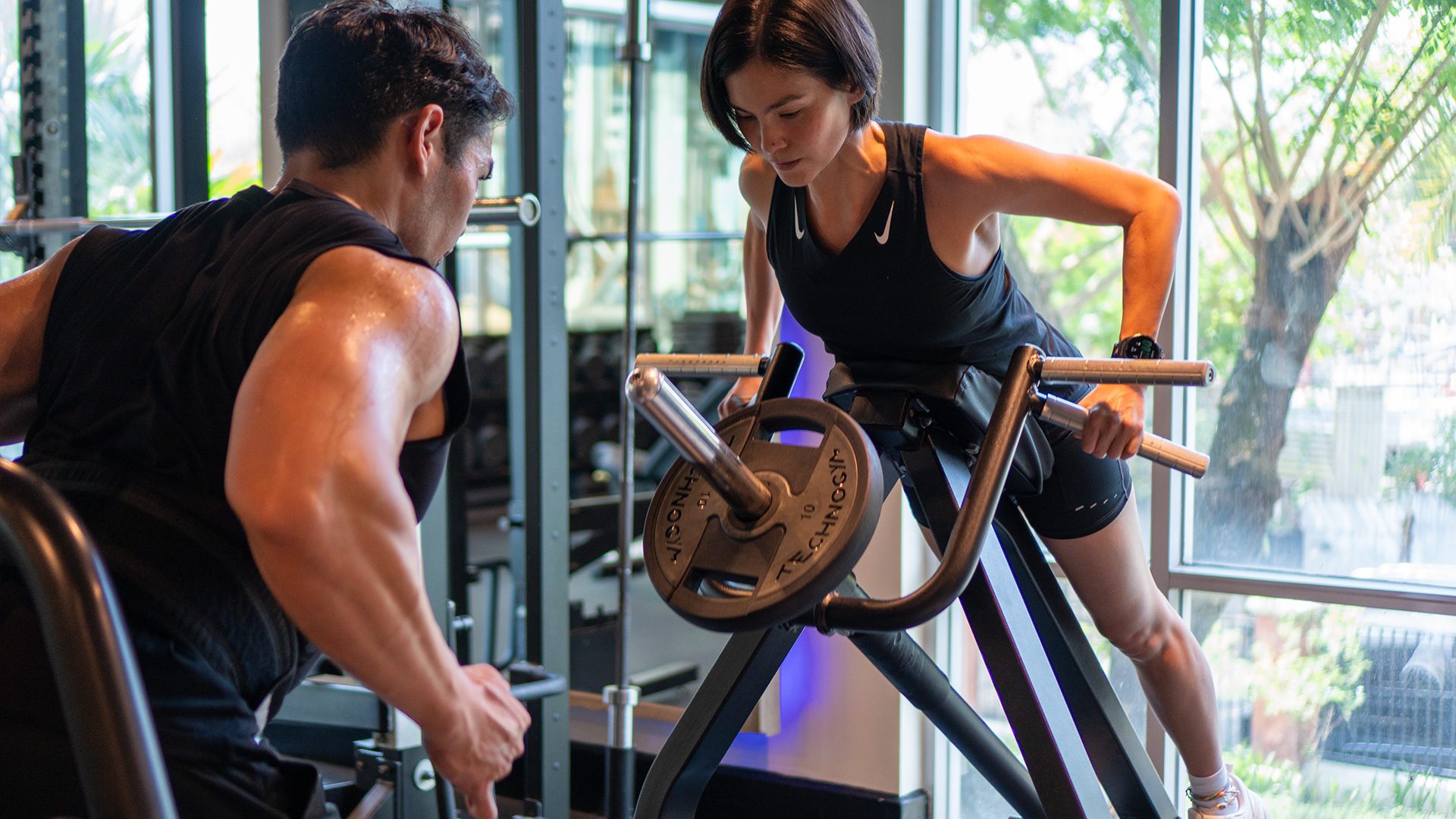 A coach spotting a member mid-set on the incline lever row at Rockwell Atletica, daylight coming through the full-height windows.
