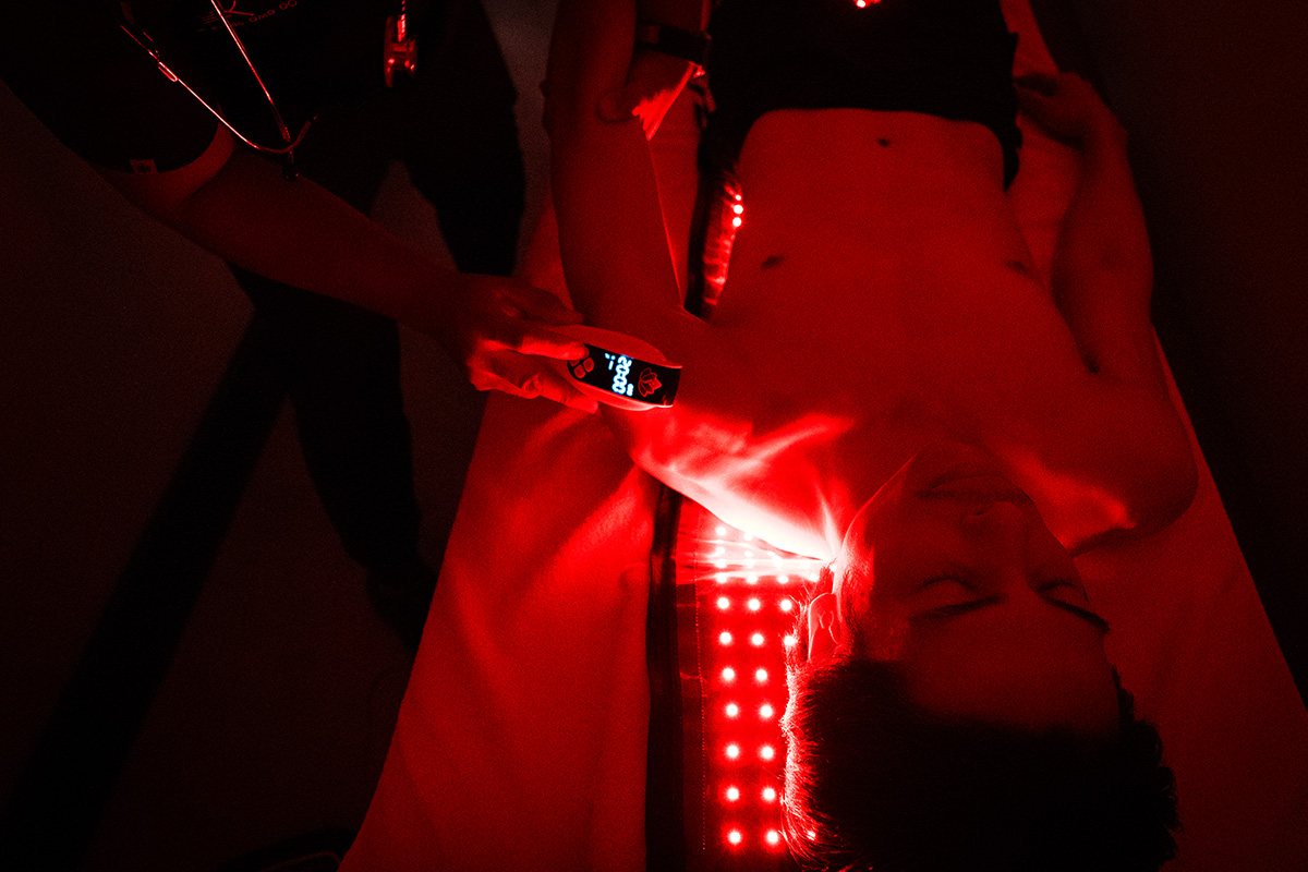 A member lying under a red light therapy panel while a recovery team member adjusts the session.