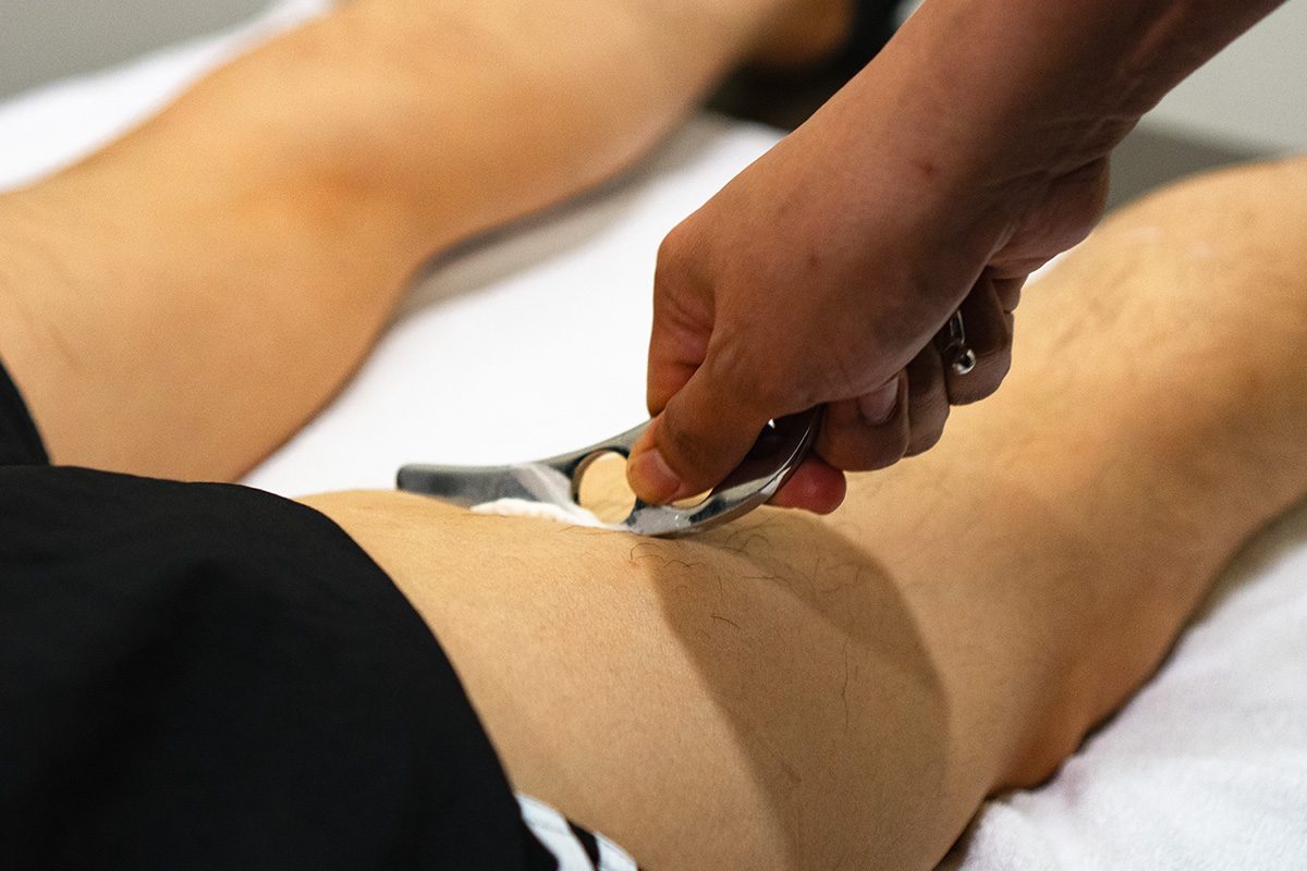 A recovery team member working a member's leg with a stainless blade tool on the treatment table.
